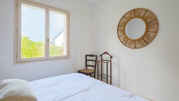Un dormitorio con cama, ventana, una silla y un espejo fijado a la pared.