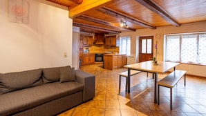 A living room with a sofa, a wooden dining table with benches, a kitchen with wooden cabinets, and a tiled floor.