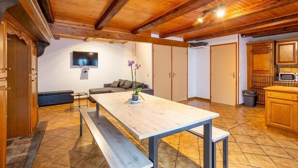 A room with a wooden ceiling, a dining table with benches, a sofa, a TV mounted on the wall, and a kitchen area with cabinets and a microwave.