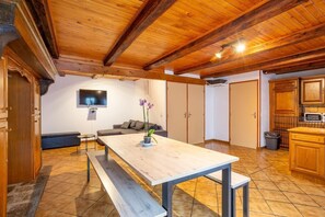 Quarto com teto de madeira, mesa de jantar com bancos, sofá, televisão na parede e cozinha com armários e micro-ondas.