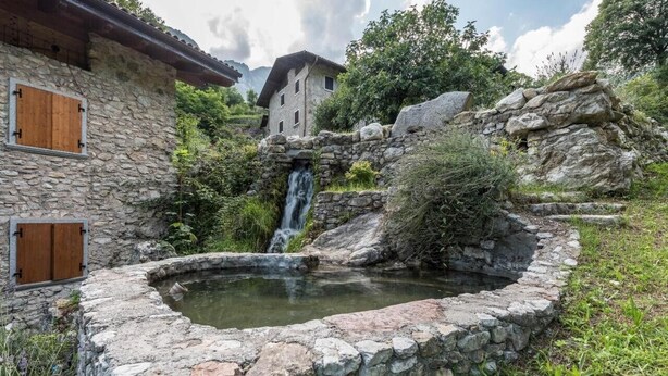 Een rustiek stenen gebouw met een kleine waterval en vijver, omringd door groen en bergen.