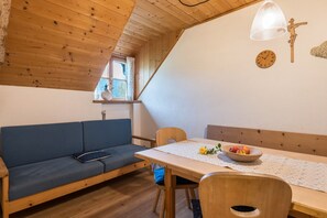 A cozy room with a wooden ceiling, a blue sofa, a wooden dining table with a bowl of fruit, and a wall clock.