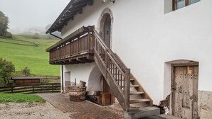 Un edificio rústico con una escalera de madera, un barril de piedra y una puerta de madera.