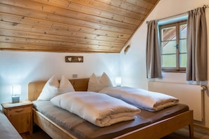 Quarto com teto de madeira, cama com roupa de cama branca, mesa de cabeceira com abajur e janela com cortinas.