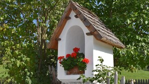 Una pequeña estructura blanca con techo a dos aguas, rodeada de follaje y una cerca de madera.