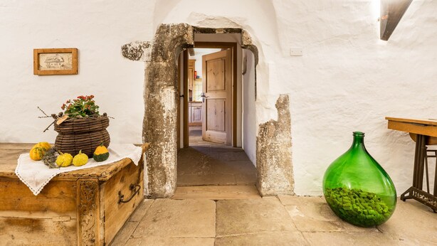 Una habitación rústica con un cofre de madera, un jarrón de vidrio verde y una puerta que da a otra habitación.