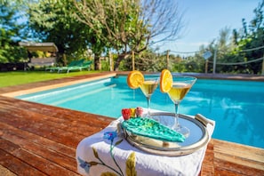 A poolside setting with a table holding a martini glass and a cocktail, surrounded by greenery and a clear blue pool.