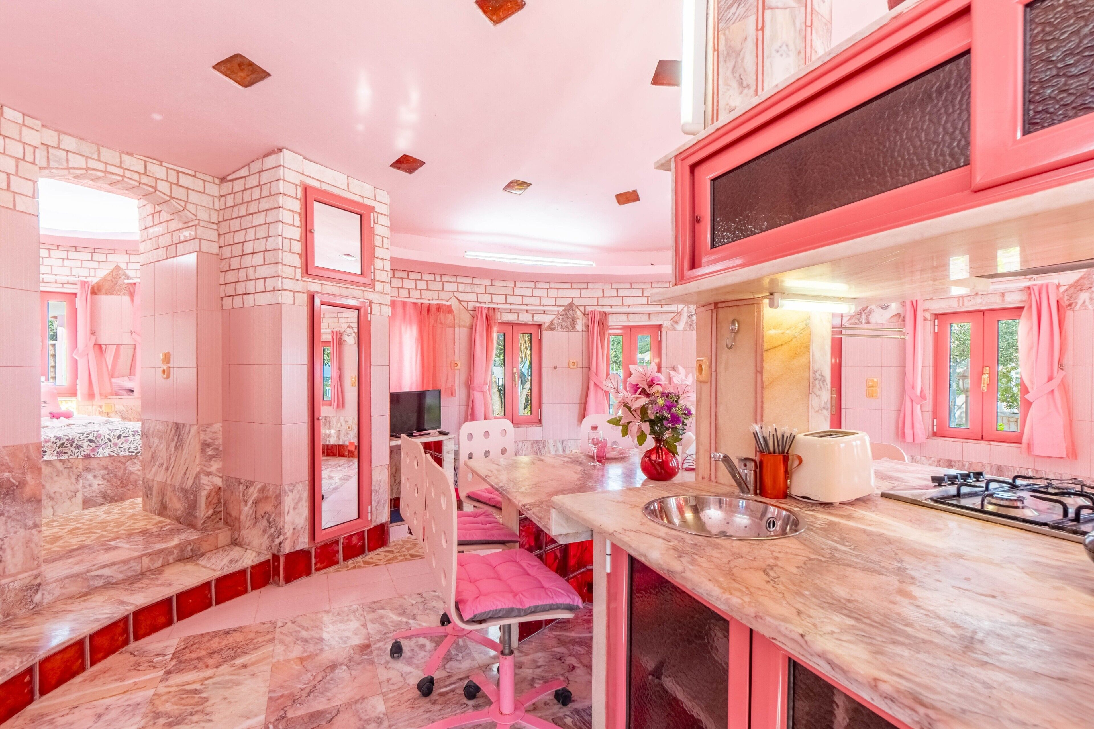 A pink-themed kitchen with a marble countertop, a sink, a stove, and a dining area with a chair and table.