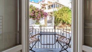 An open door leading to a balcony with a table and chairs, a view of a white building with a sign, and a flowering bush. - Studio Apartment 'En Tino 1' with Balcony, Wi-Fi and Air Conditioning (Tinos)