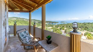 Un balcone con soffitto in legno, una sedia con cuscino floreale, un tavolino con una pianta in vaso e vista sul paesaggio costiero.
