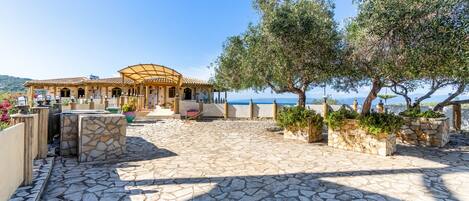 A stone-paved patio with outdoor seating, a pergola, and olive trees.