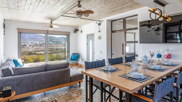 A modern dining area with a large table set for six, a sofa, and a view of a mountainous landscape through large windows. - Country House 'A The' with Mountain View, Shared Terrace and Wi-Fi (Tinos)