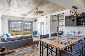 A modern dining area with a large table set for six, a sofa, and a view of a mountainous landscape through large windows.