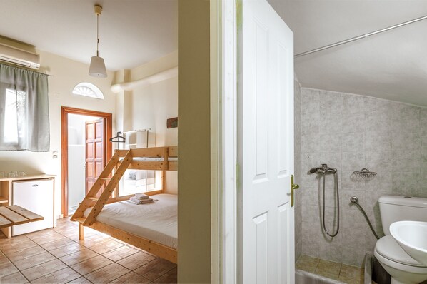 A room with a bunk bed, a small desk, a window with curtains, and a bathroom with a shower and toilet.