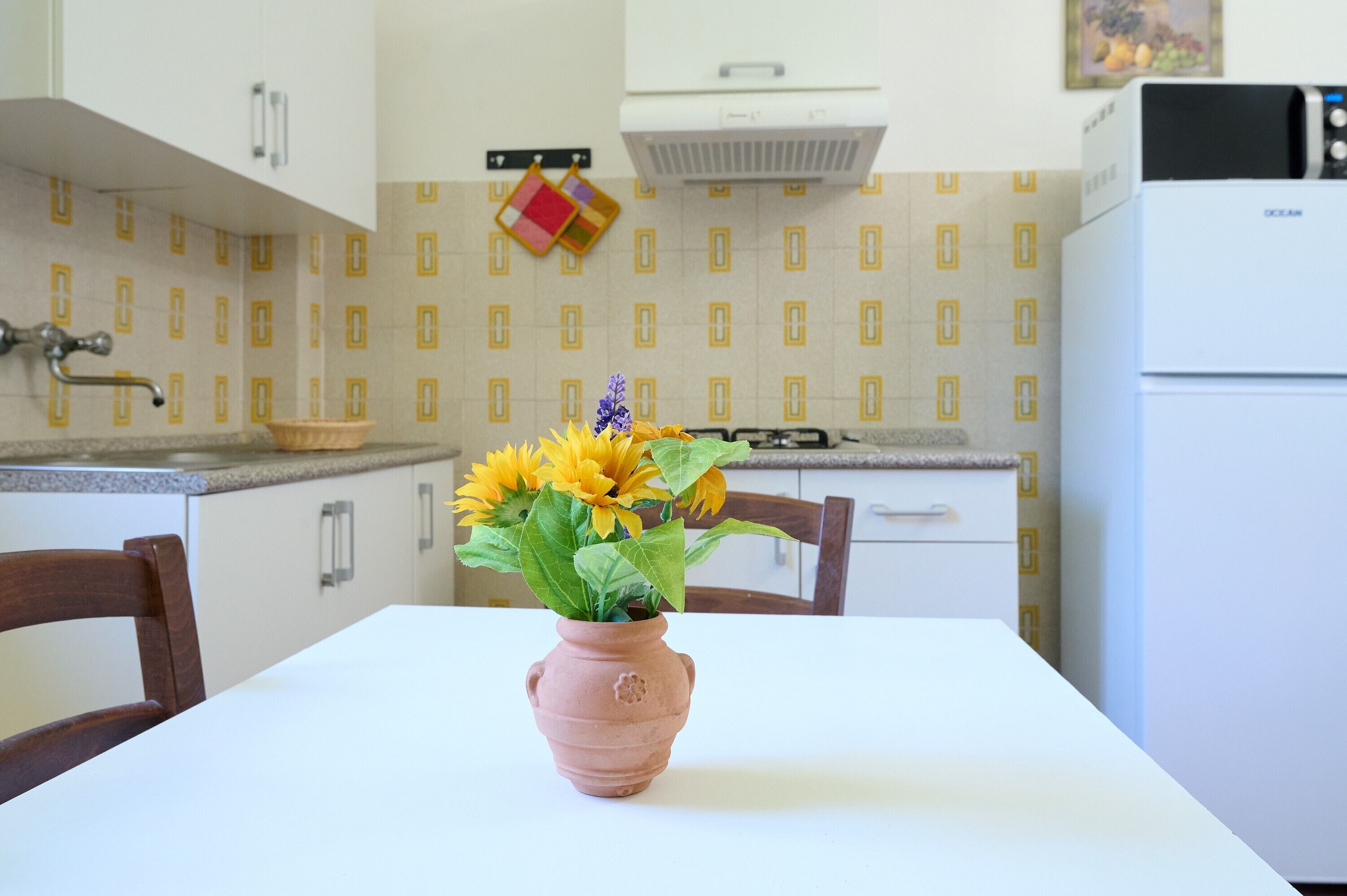 Een keuken met een witte tafel, een roze vaas met bloemen en een decoratieve spatwand.