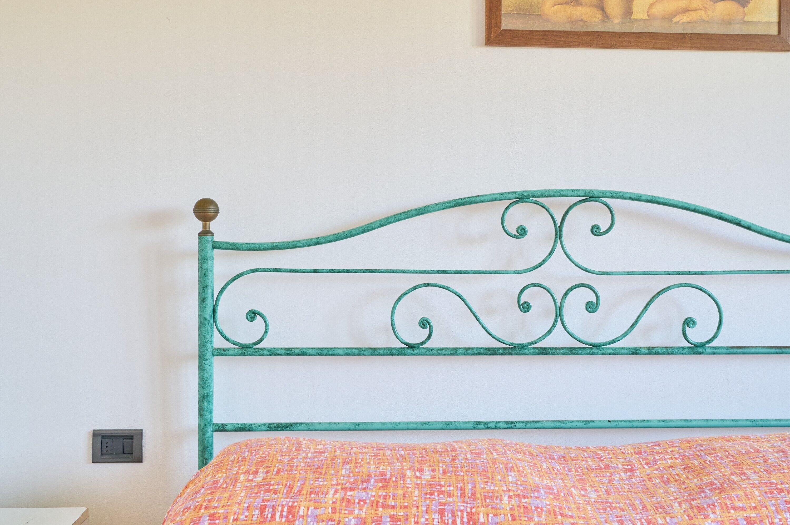 Un lit avec une tête de lit en métal vert, une couvre-lit à motifs orange et rose, et une prise électrique au mur.