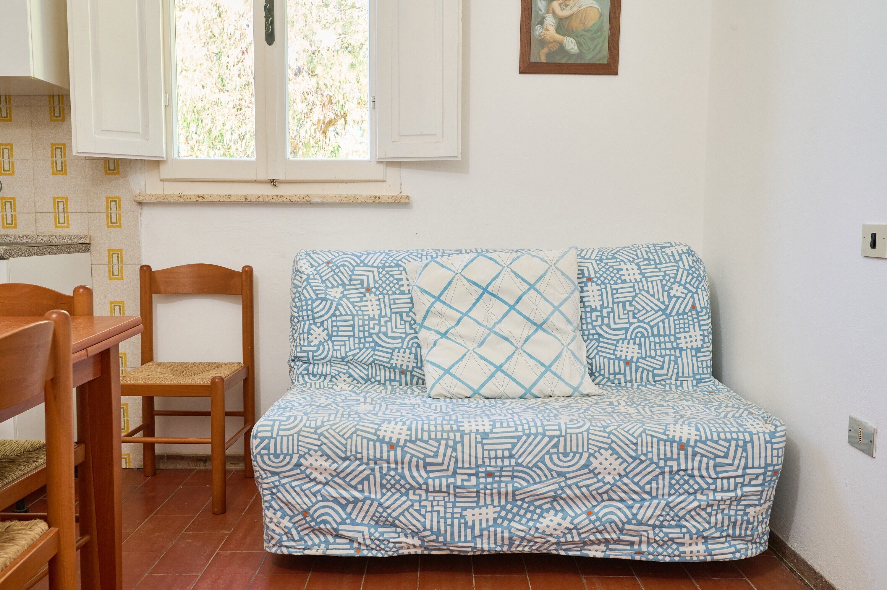 A room with a patterned sofa, a wooden chair, a dining table, and a window with shutters.