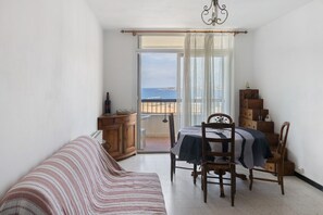 A bedroom with a bed, a dining table with chairs, a wooden dresser, and a view of the sea through a large window.