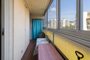 Ein schmaler Gang mit einem Bett auf der rechten Seite, einem blauen Vorhang und einem Fenster mit Blick auf Gebäude.