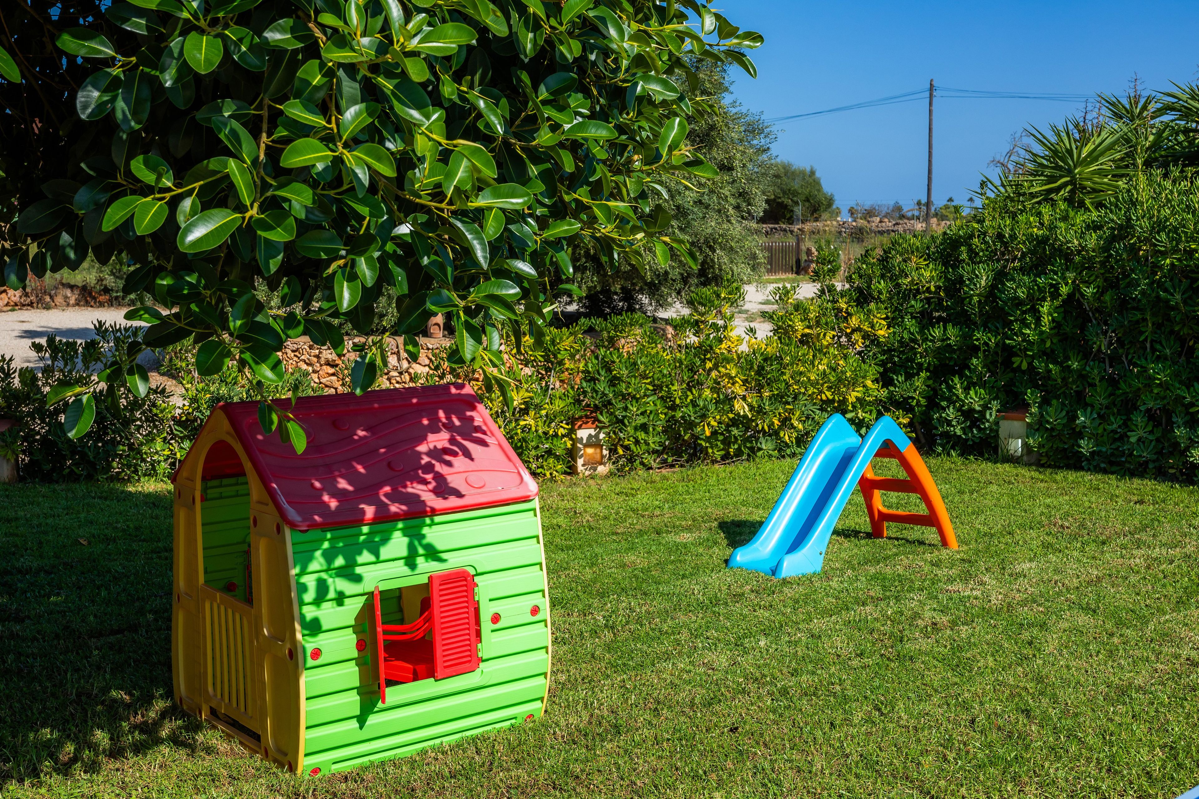 A children's playhouse with a red roof and green walls, a blue slide, and a grassy yard with a large tree and bushes.