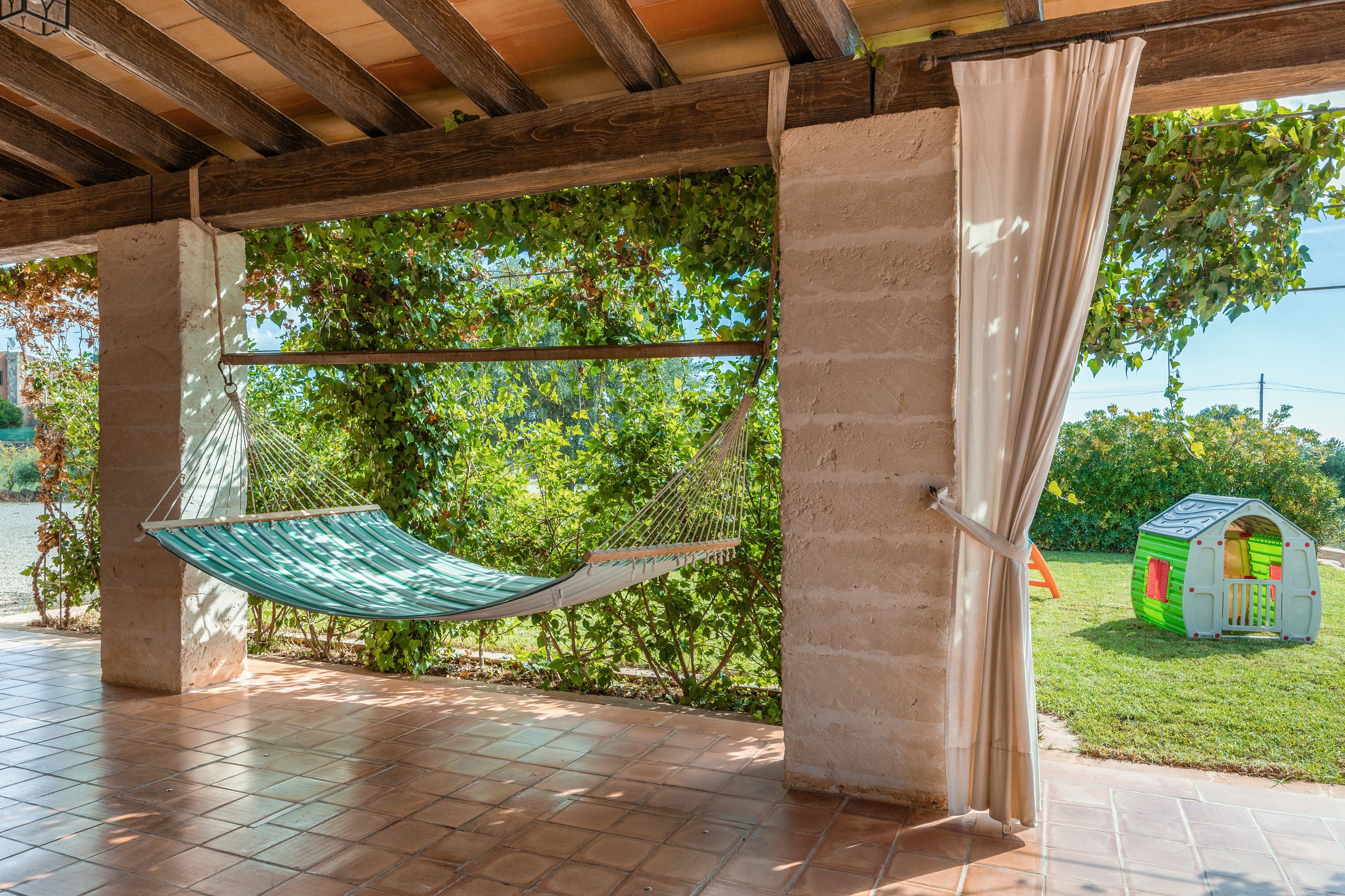 A hammock under a wooden pergola with a view of a green playhouse.