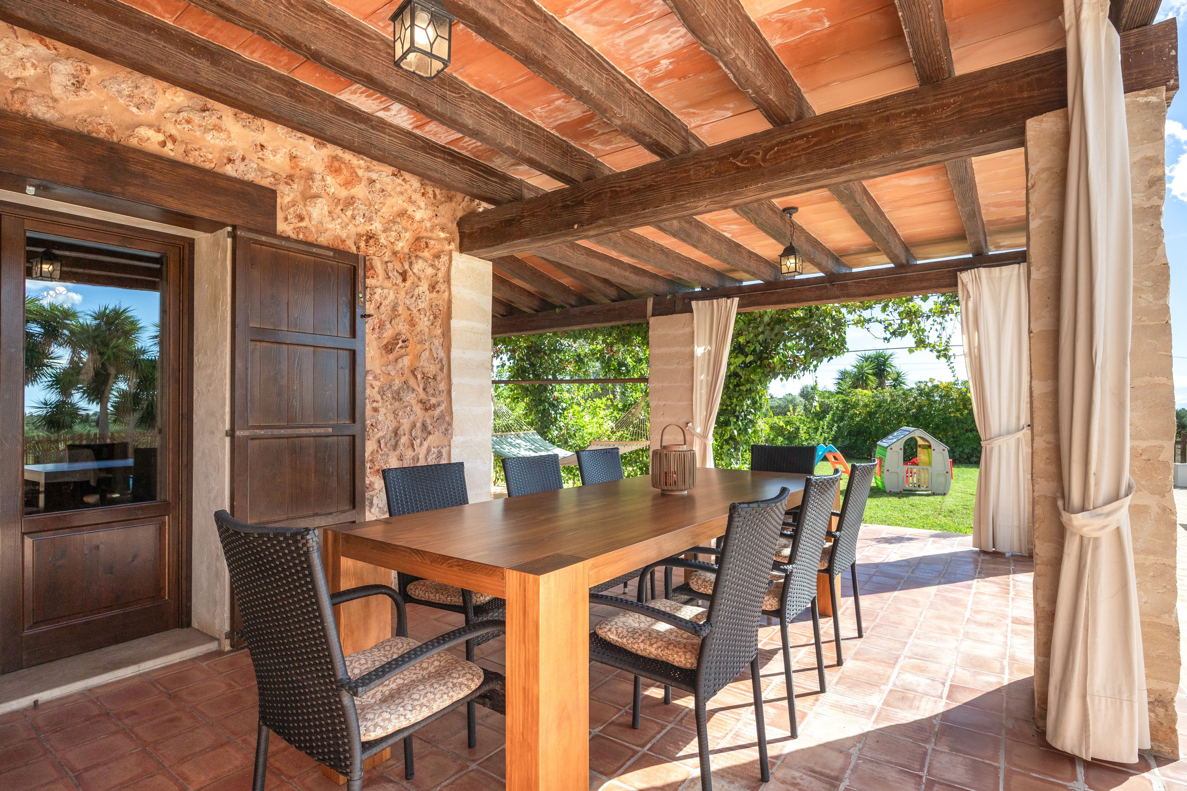An outdoor patio with a wooden table and chairs, stone walls, and a view of greenery.