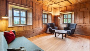 Een kamer met houten lambrisering, een bank, twee fauteuils, een ronde tafel en twee ramen.