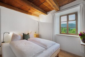 A bedroom with a wooden ceiling, a large window with a view of greenery, a bed with white bedding, and a bedside table with a lamp. - Apartment 'Ansitz Baumannhof 1818' with Mountain View, Shared Garden and Wi-Fi (Pfalzen)