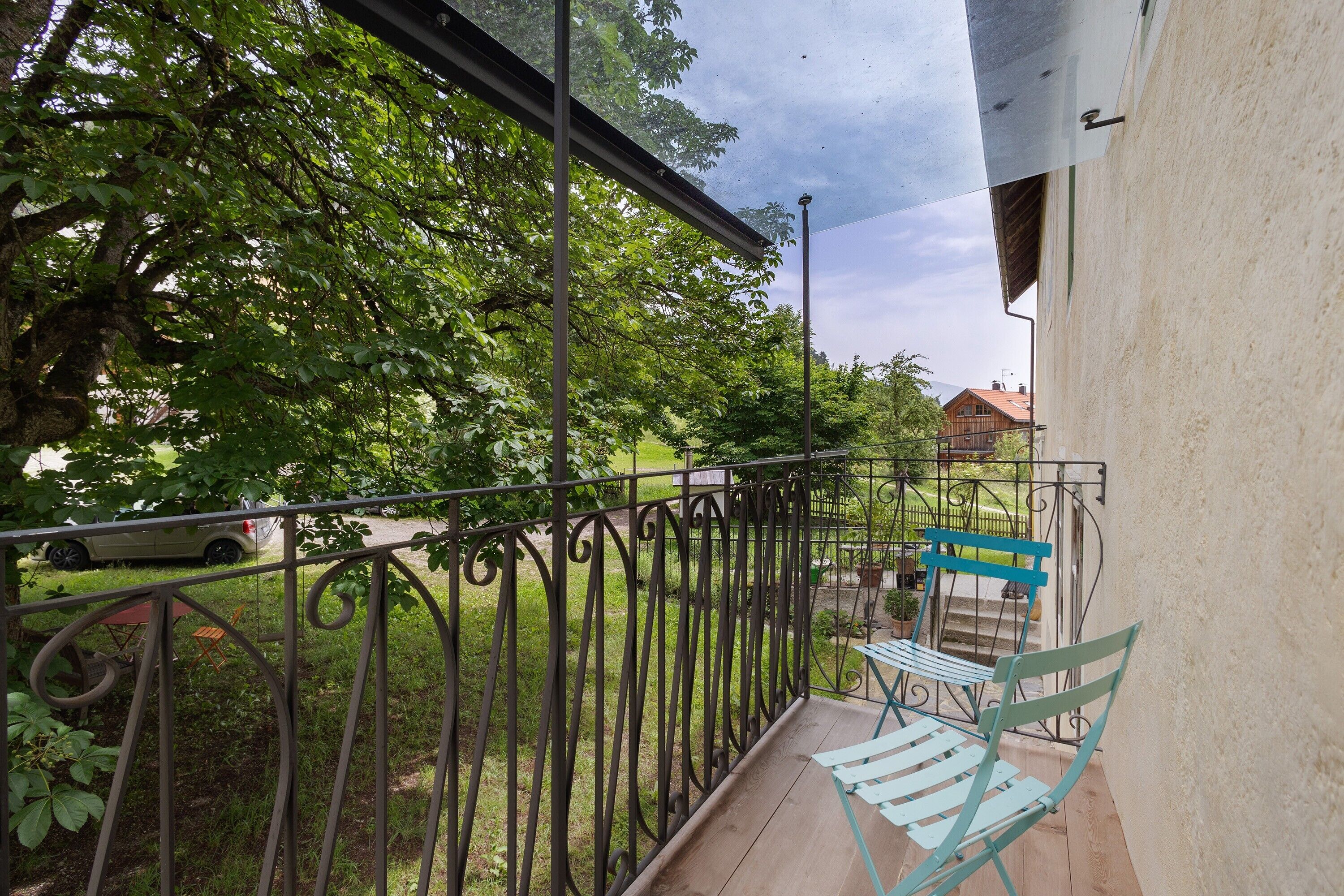 Un balcon doté d’une balustrade en métal, d’un plancher en bois et offrant une vue sur un paysage verdoyant, avec des arbres et un bâtiment situé au loin.