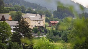 A large building with multiple windows and a sloped roof, surrounded by lush greenery and hills.