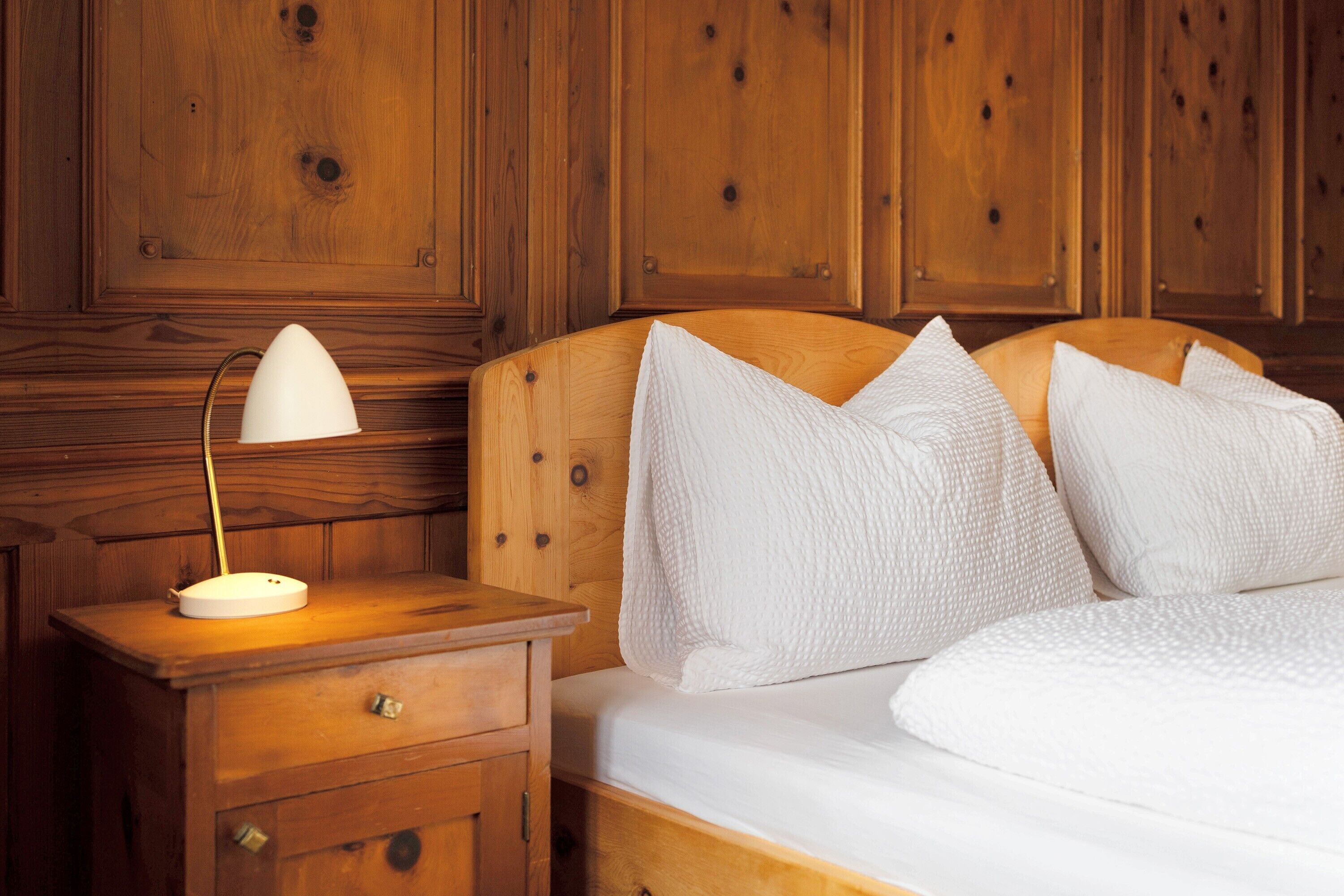 Une tête de lit en bois, agrémentée de coussins blancs, une lampe de chevet et une table de chevet en bois avec un tiroir.