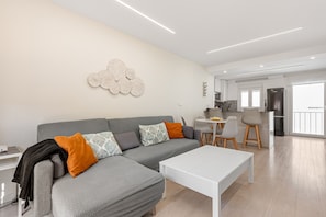 A modern living room with a grey sofa, white coffee table, and a kitchen in the background.