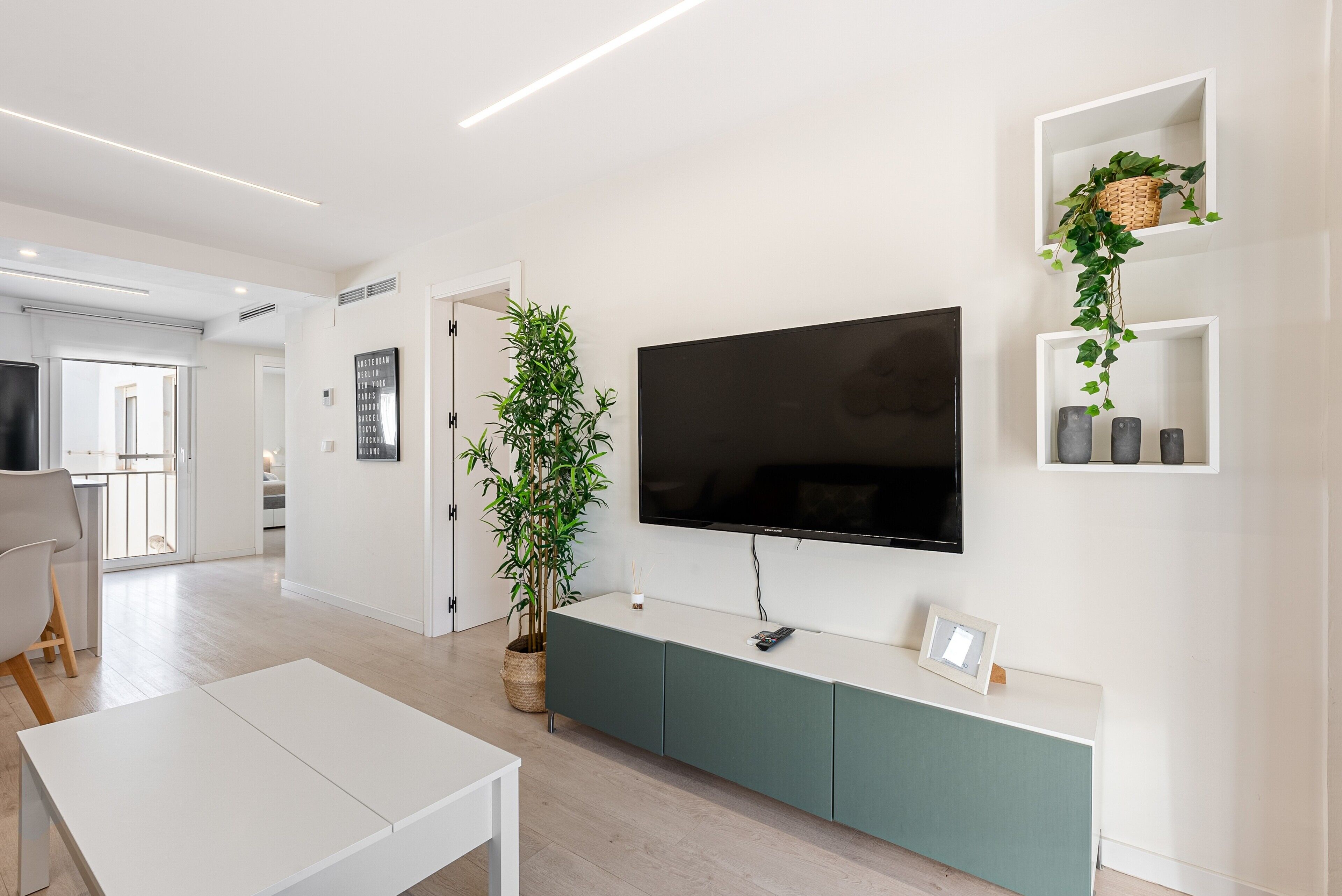 Un salón moderno con televisor de pantalla plana, una consola verde, una planta en maceta y una mesa de centro blanca.