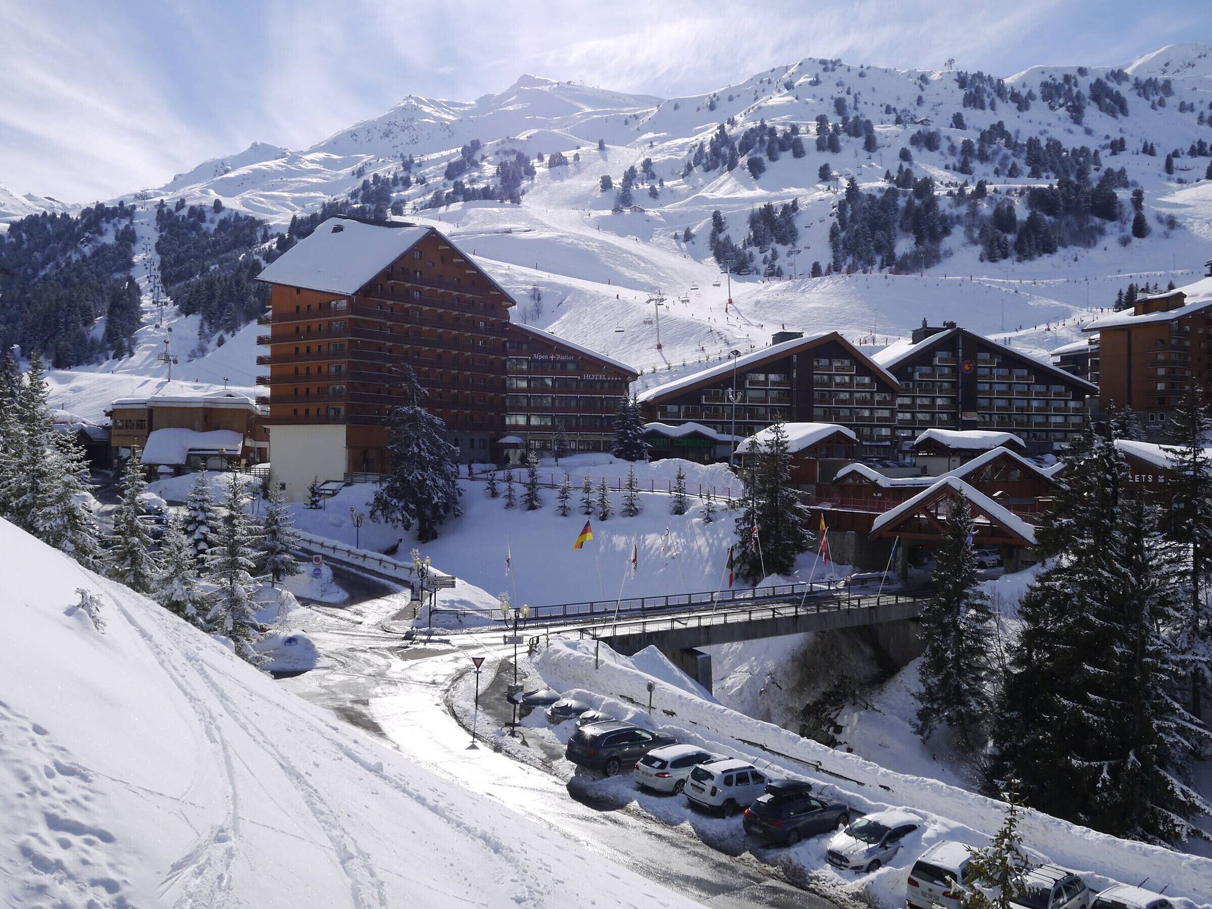 A snow-covered mountain resort with multiple wooden buildings, ski lifts, and a parking area.