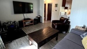A living room with a flat-screen TV, a wooden coffee table, a sofa, and a kitchen area in the background. - Puma's executive cat house! (Fernie)