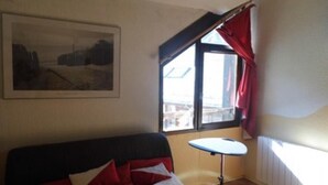 A room with a bed, a small table, a window with curtains, and a painting on the wall. - Apartment 'Azur Sancy Skis Aux Pieds' (Le Mont-Dore)