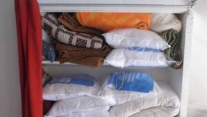 A storage closet with shelves holding bags of various items, a red curtain, and a wooden bench. - Apartment 'Azur Sancy Skis Aux Pieds' (Le Mont-Dore)