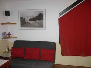 Une pièce avec un lit, un tableau encadré au mur, un rideau rouge et une étagère avec des livres et une petite statue.