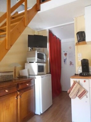Une cuisine compacte avec des meubles en bois, un réfrigérateur, un four à micro-ondes, une machine à café et un téléviseur posé sur une étagère.