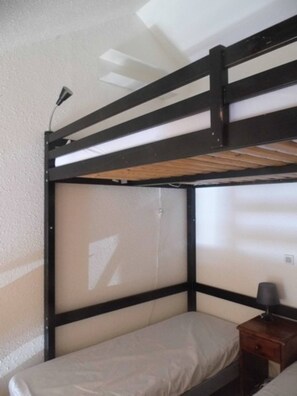Una cama litera con estructura de madera y una cama individual debajo. Hay una mesita de noche con una lámpara y un cajón.