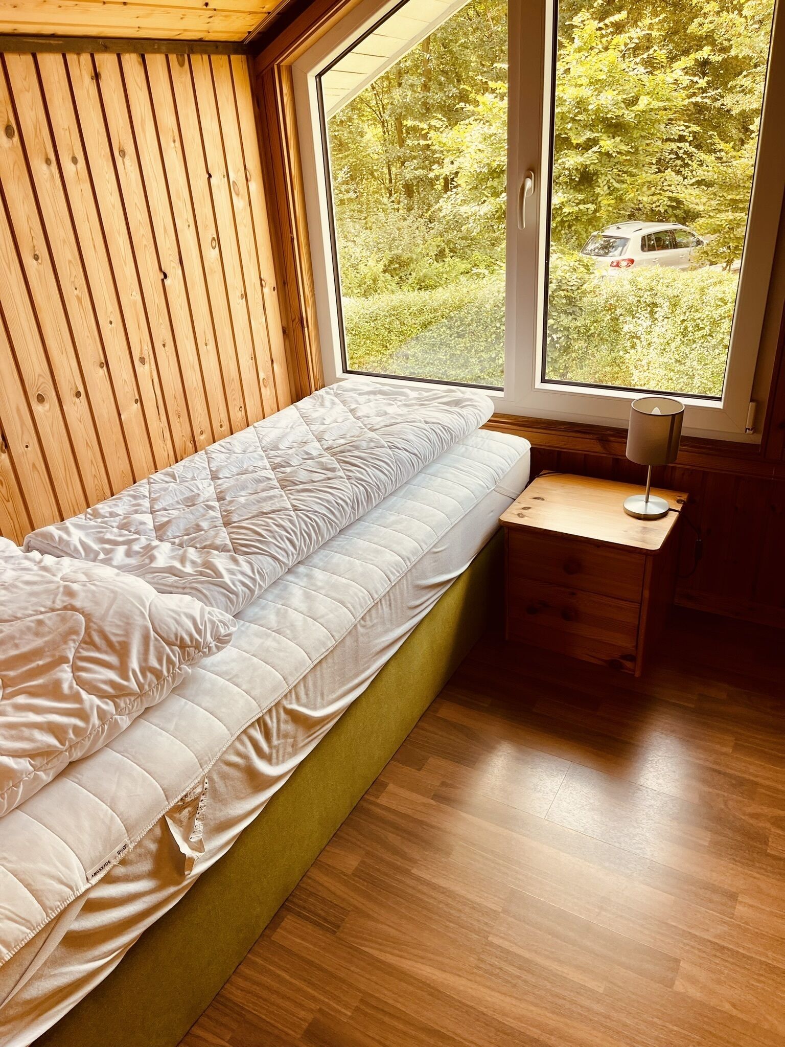 A bedroom with a bed against a wooden wall, a window with a view of greenery, and a bedside table with a lamp.