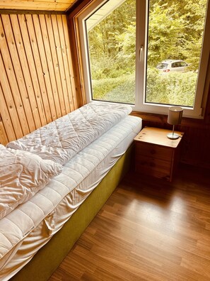 A bedroom with a bed against a wooden wall, a window with a view of greenery, and a bedside table with a lamp. - Premium-ferienhaus im Extertal Ferienpark - Wandern, Natur, Wald, Sauna # (Extertal)