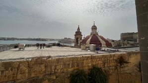A coastal view with a domed building and two towers, a paved area with tables and chairs, and a stone wall in the foreground. - Valletta heart, 3-room superior apartment (Valletta)