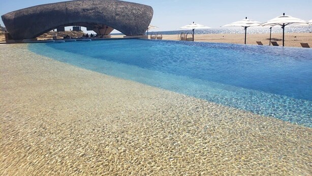 A clear pool with a unique rock arch structure, a sandy beach, and umbrellas.