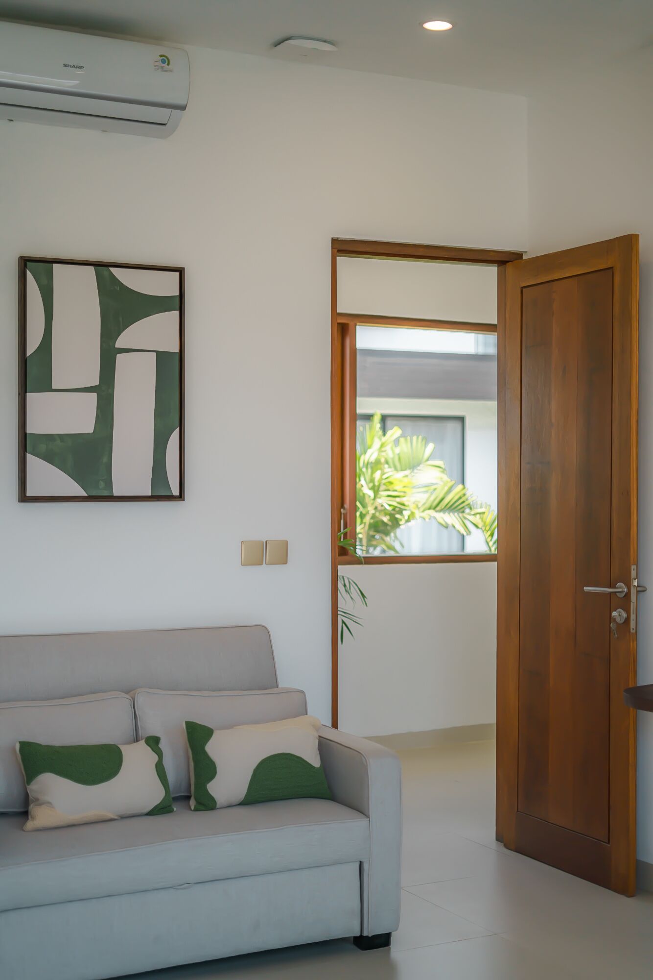 En moderne stue med en grå sofa, et abstrakt maleri i grønne og hvide farver, samt en trædør der fører ud til en balkon med planter.