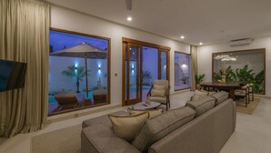 A modern living room with a large sofa, a coffee table, and a TV. There are large windows with views of a pool and palm trees. - Villa Umaju  (Kerobokan)