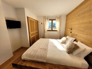 A bedroom with a wooden headboard, a bed with white linens, a wooden floor, a TV mounted on the wall, and a window with curtains. - Bright apartment for 6 with view and parking (Les Gets)
