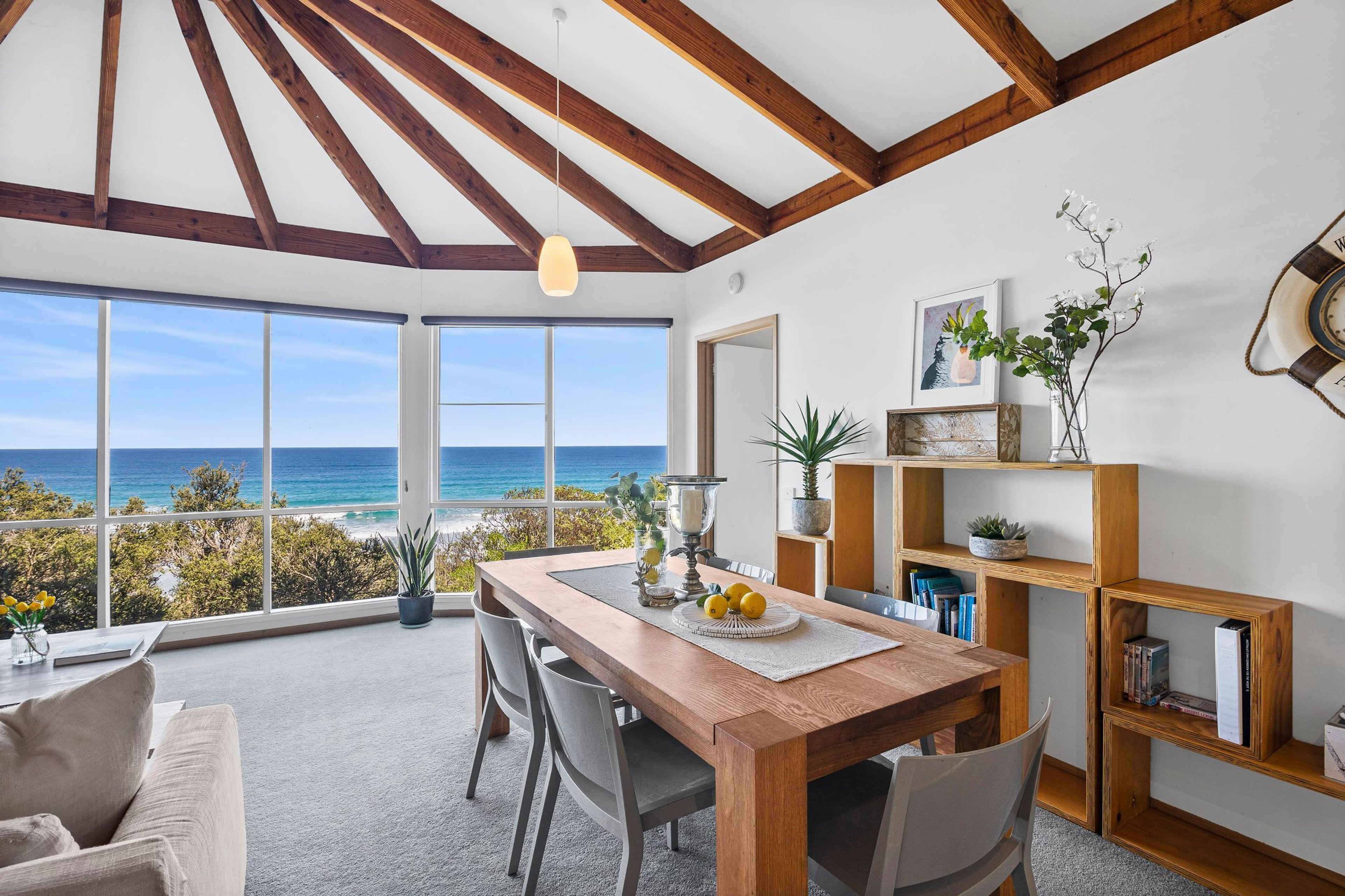 A dining area with a view of the ocean, wooden beams on the ceiling, and a bookshelf with books and plants.