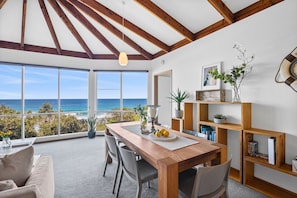 Un comedor con vistas al océano, vigas de madera en el techo y una estantería con libros y plantas.