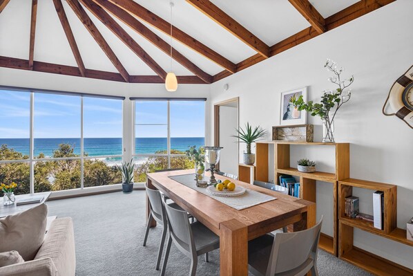 A dining area with a view of the ocean, wooden beams on the ceiling, and a bookshelf with books and plants.
